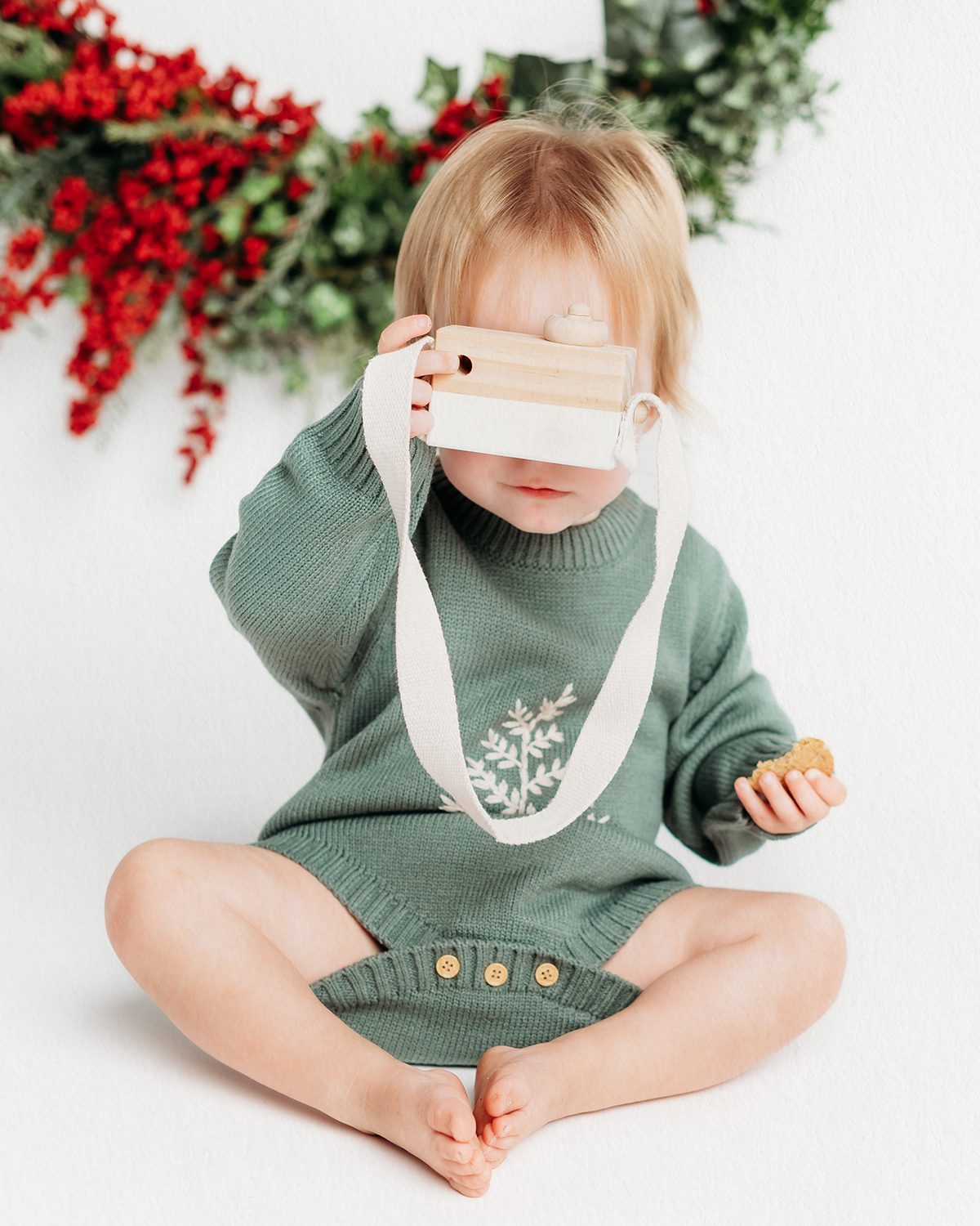 a baby girl holding up a toy camera to her eye during a themed christmas mini photo shoot