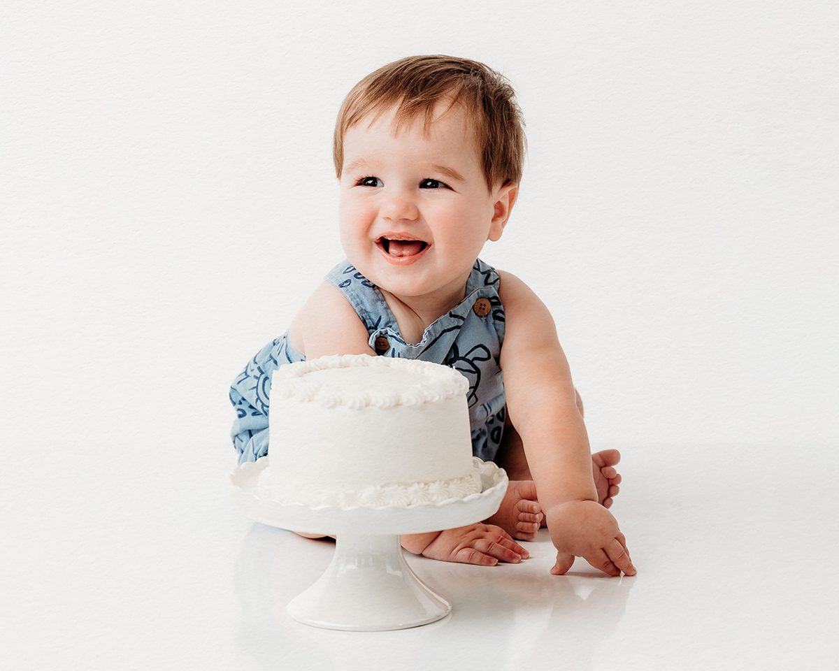 baby first birthday a baby boy and a birthday cake set against a plain white backdrop during a first birthday photoshoot