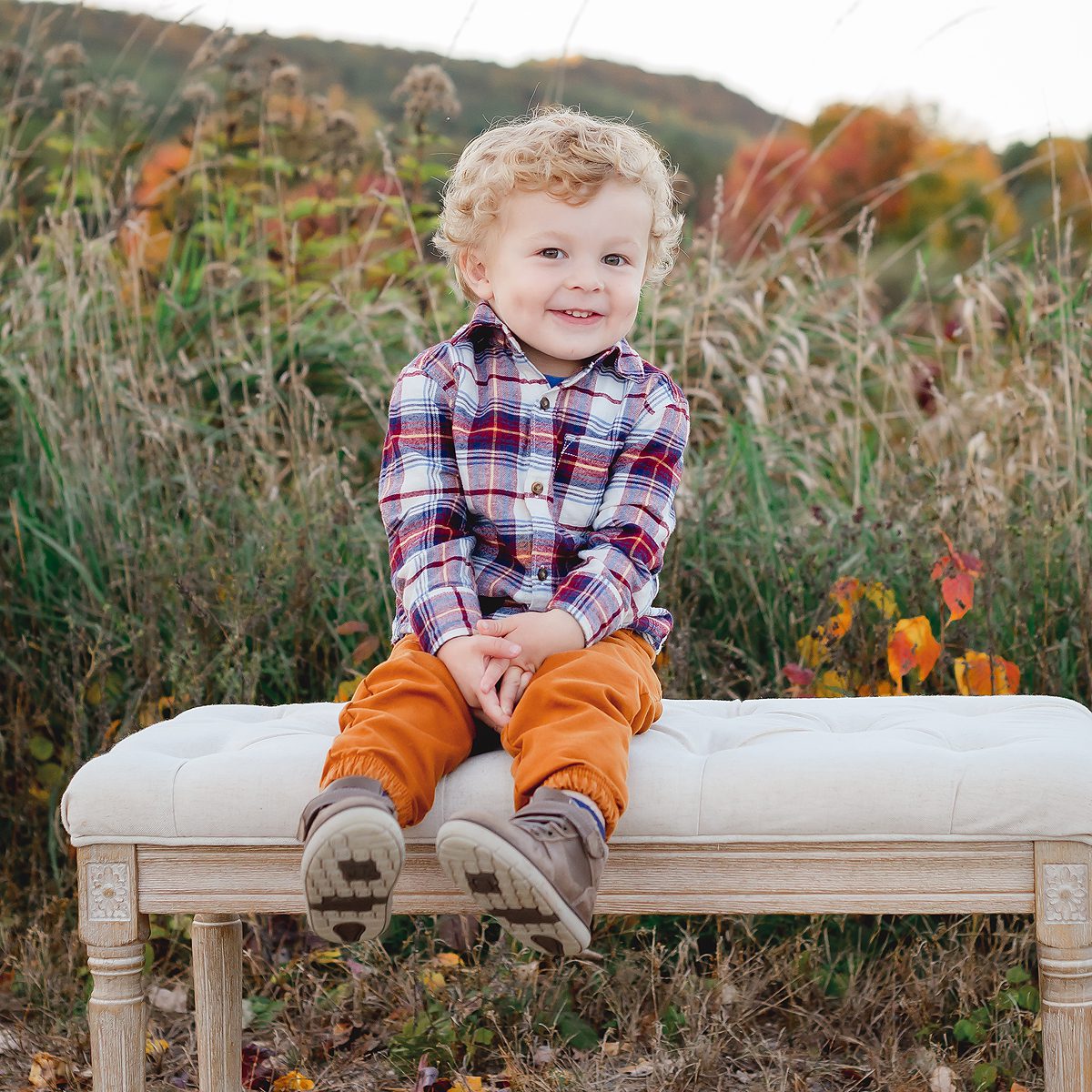 003_CT_OUTDOOR_FALL_MINI_SESSIONS A little boy wearing orange pants and a plaid shirt, sitting on a bench in a field of tall grasses during autumn in Connecticut. He is smiling at family photographer Kelli Dease.