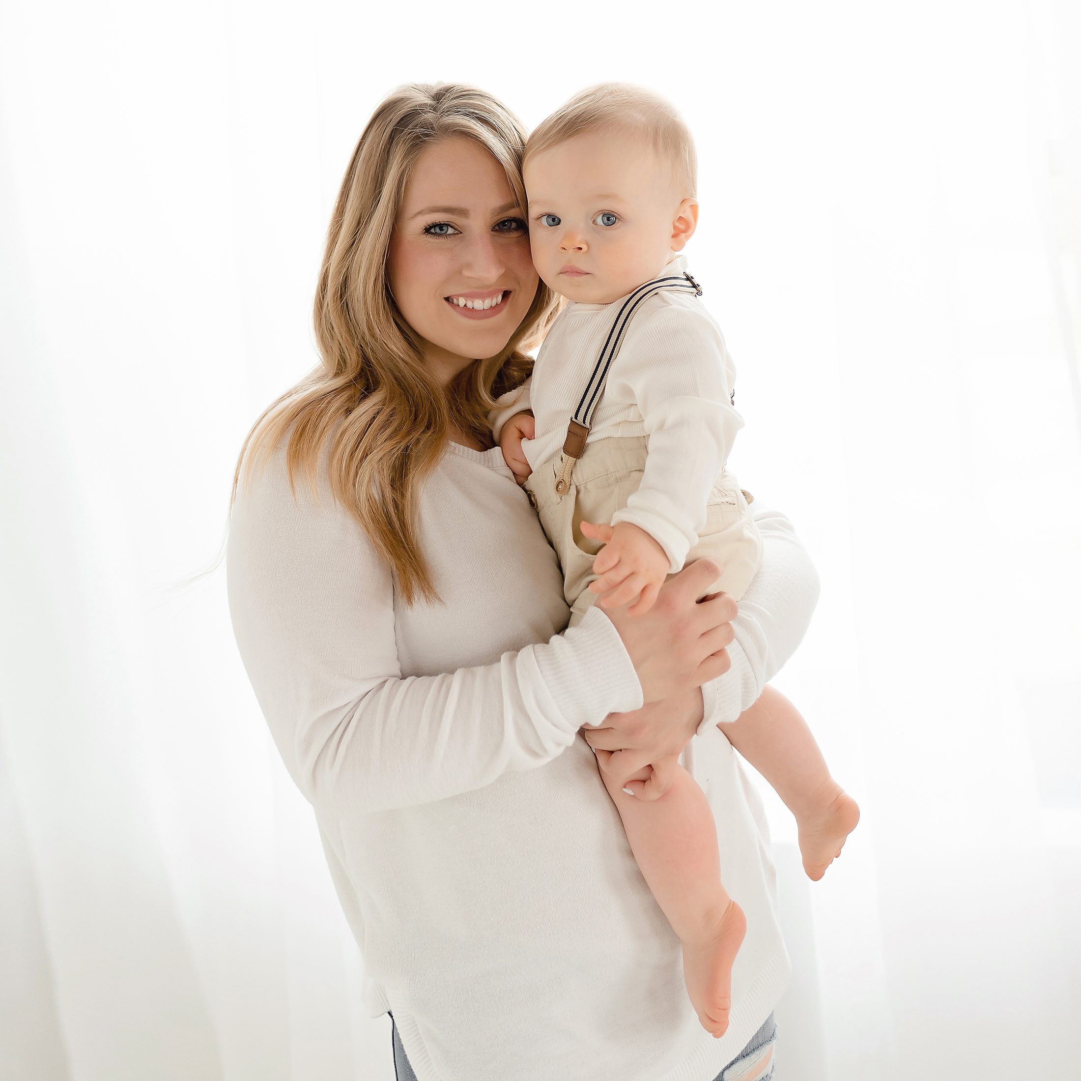 15_CT_SIMPLE_CAKE_SMASH_PHOTOGRAPHER_KELLI_DEASE A portrait of a blonde mother holding her one-year-old baby boy in front of a window with sheer white curtains.