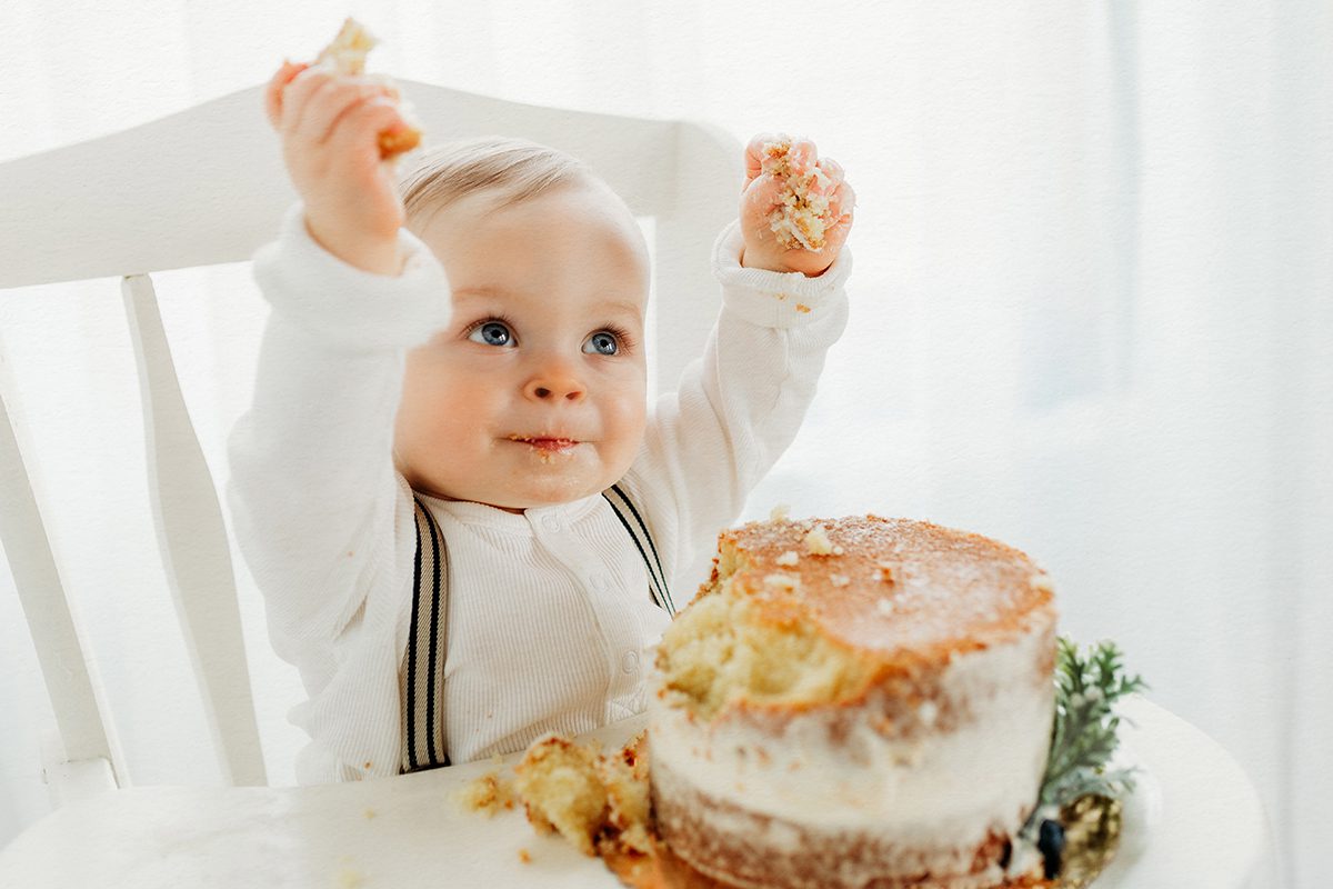 A one-year-old baby boy wearing a white Henley and suspenders, holding fists of birthday cake and smiling at the camera. The photo features a plain white background and a white high chair.