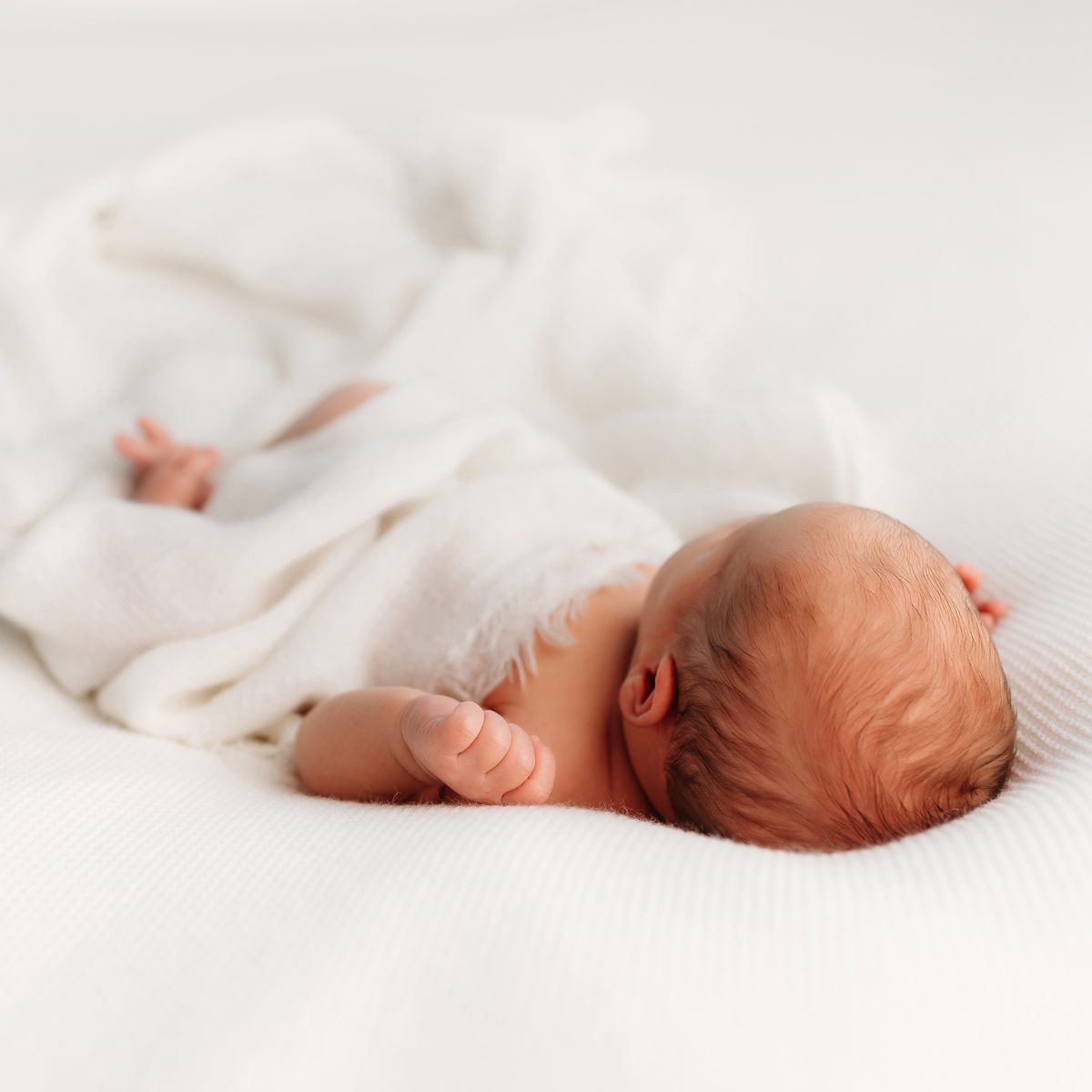 Overhead newborn photograph of a baby lying on their stomach, loosely wrapped in white fabric, photographed in a minimalist studio setting.