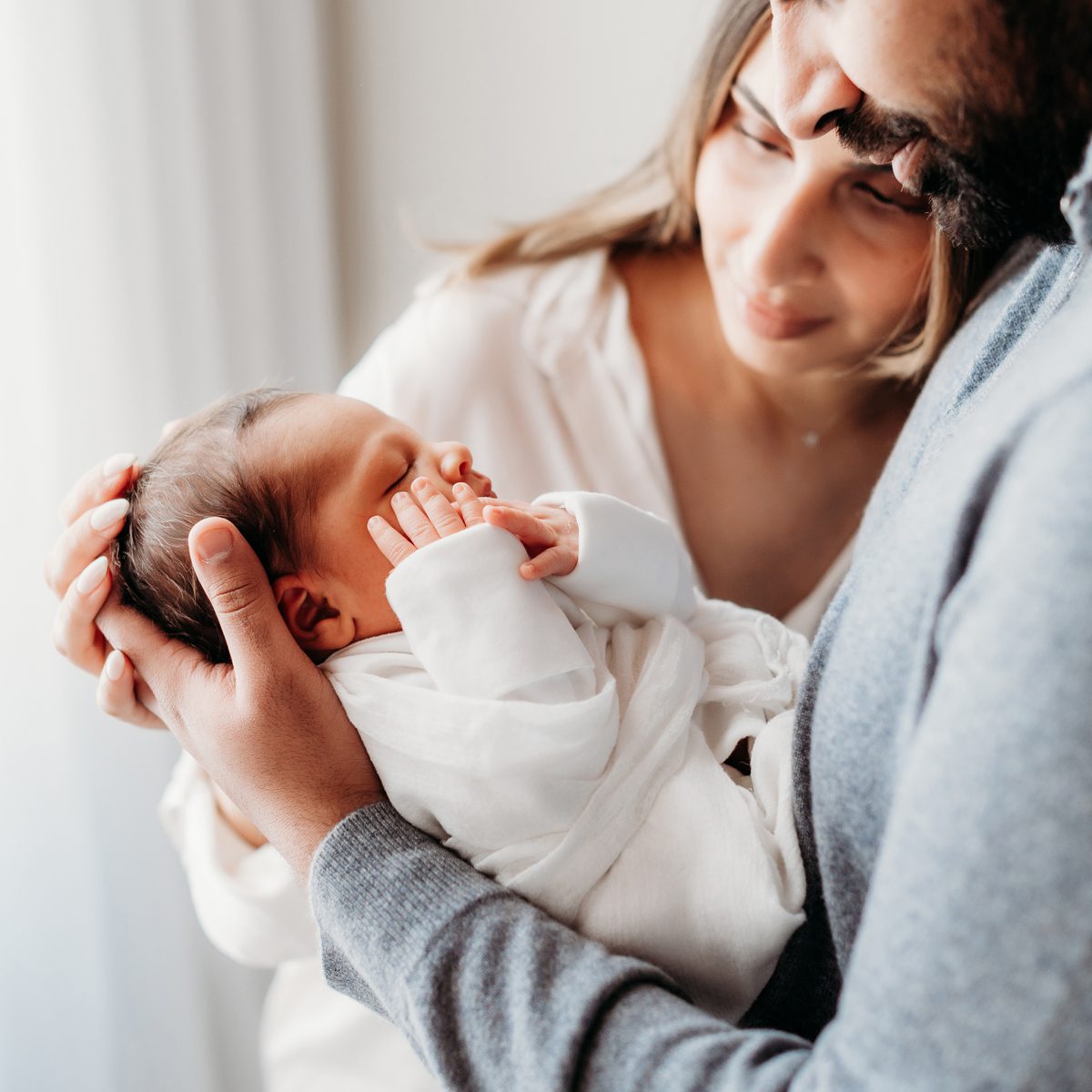 Lifestyle newborn photo of parents holding their sleeping newborn baby wrapped in white near a window, captured during a natural light newborn session with a Connecticut newborn photographer.