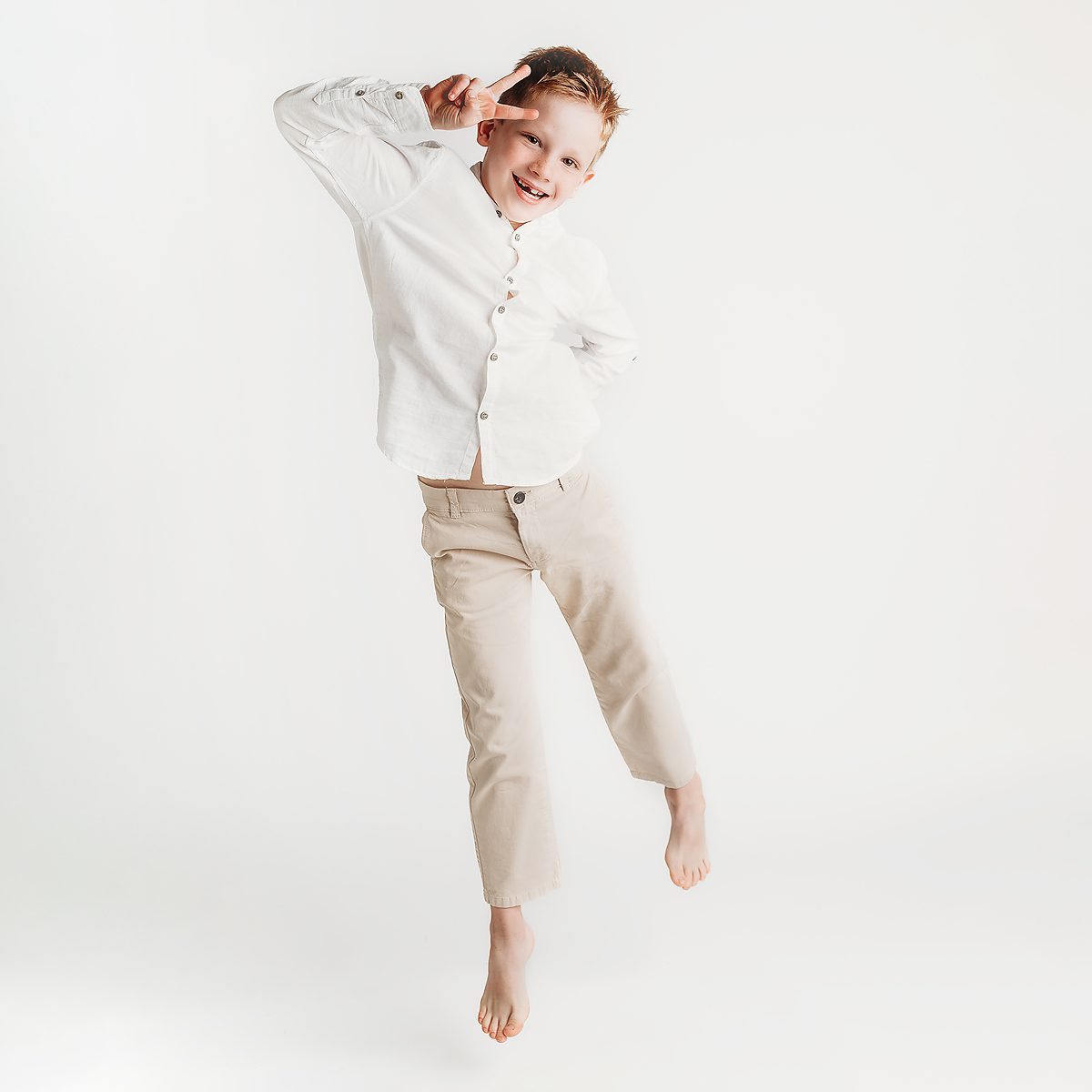 a young boy set against a white backdrop jumping and flashing a peace sign during a photoshoot