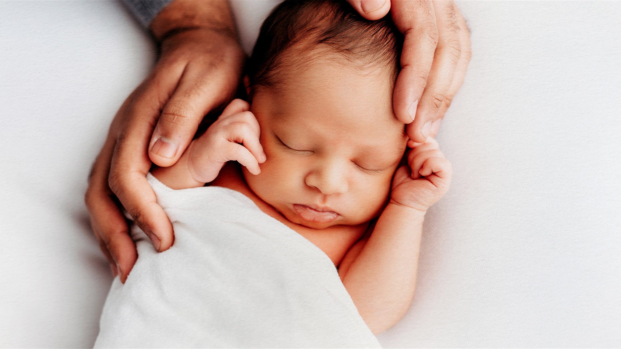 Newborn baby on white backdrop cradled by fathers hands