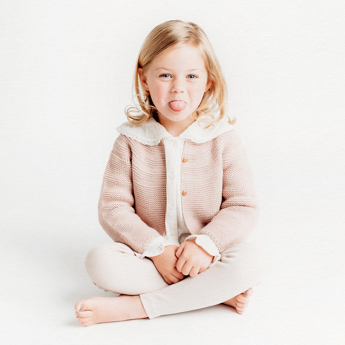 Young girl sitting cross-legged in a pink cardigan, sticking out her tongue, candid studio portrait