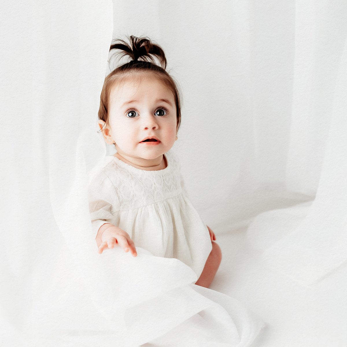 Baby girl in a cream knit dress with a dark hair top knot, seated studio portrait, white background