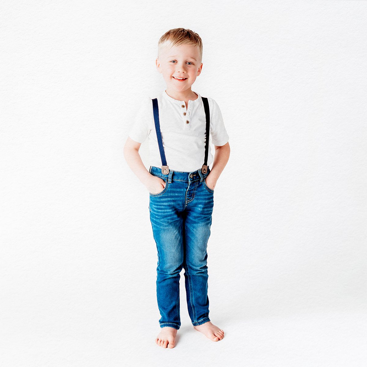 A blonde, blue-eyed little boy wearing jeans with the suspenders with his hands in his pockets, smiling at the camera during a birthday photo shoot.