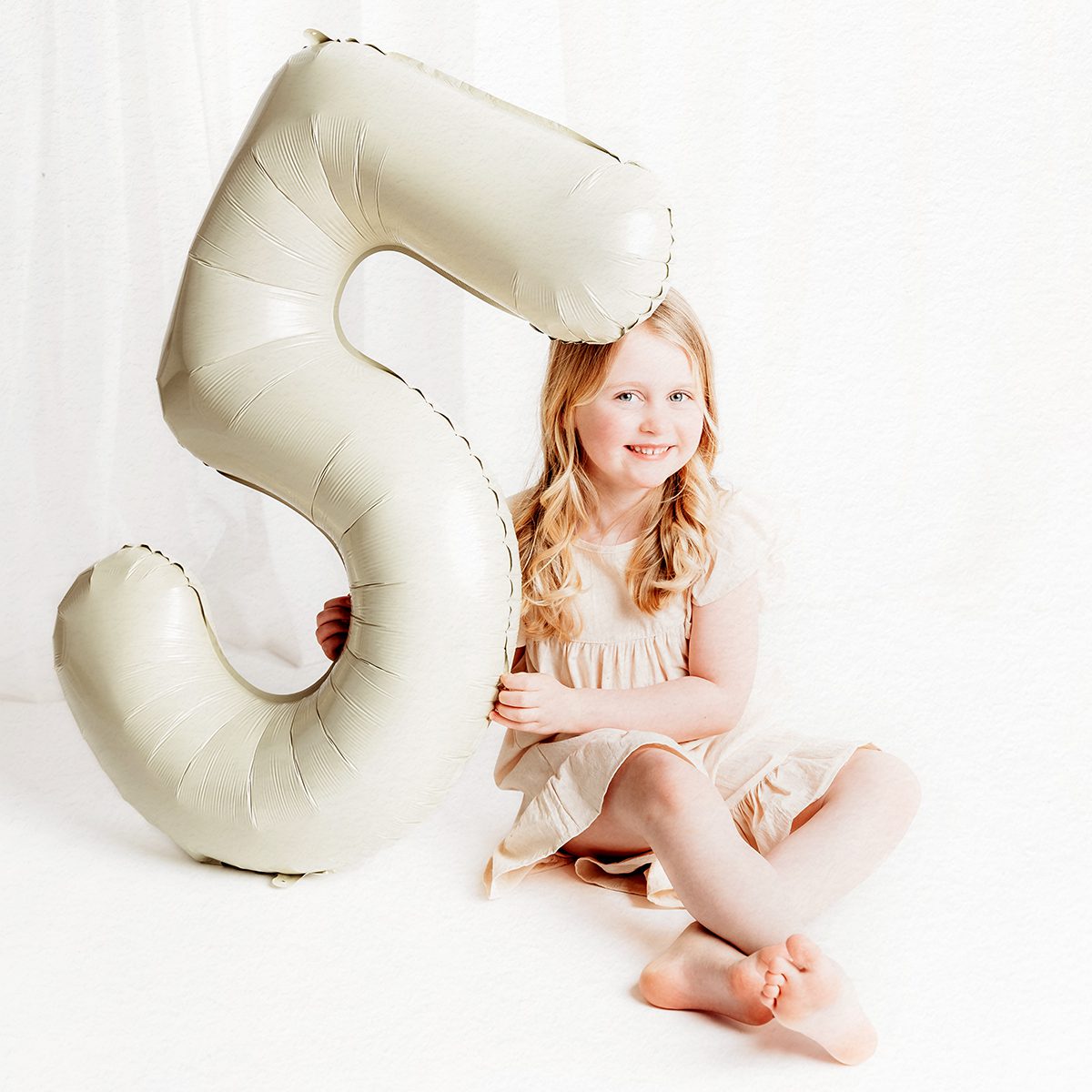 A fifth birthday portrait of a blonde girl sitting on the floor holding a number five balloon during a Farmington, Connecticut birthday photo shoot.