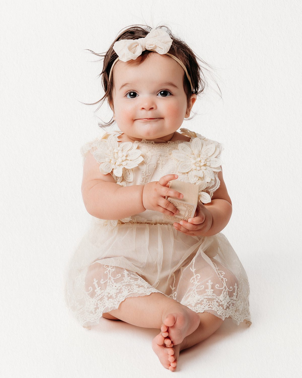 Baby girl with dark hair and a cream bow headband sitting in a cream lace dress, smiling softly while holding a wooden block during a milestone portrait session on a light backdrop.