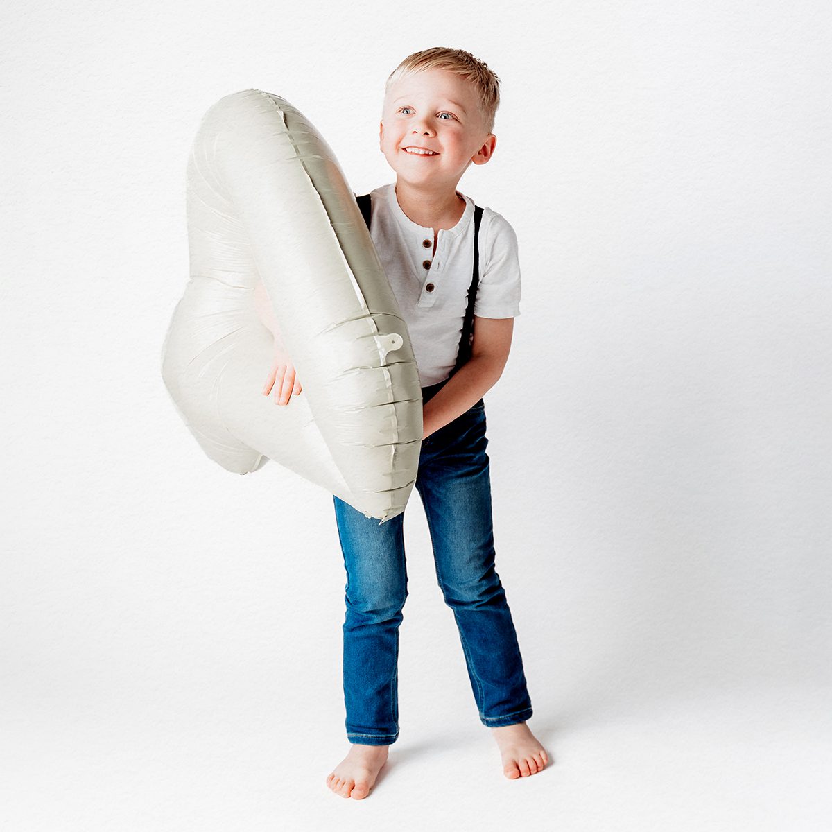 Blond boy in a white henley, navy suspenders, and jeans standing barefoot beside a large cream number 4 balloon, smiling at the camera during his 4th birthday portrait session on a light backdrop.