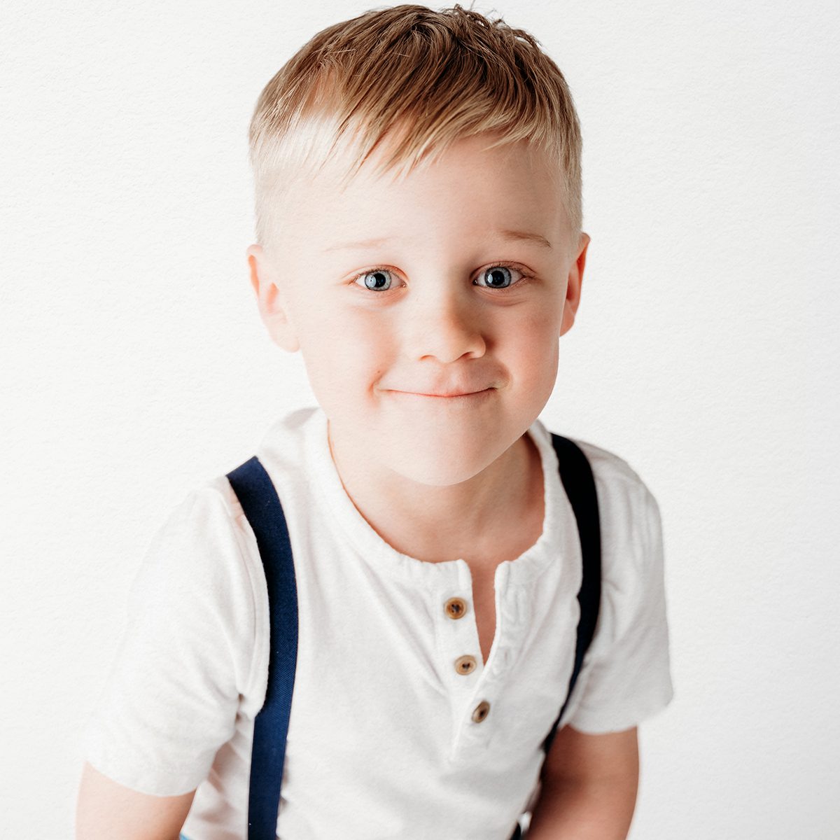 Blond boy in a white henley and navy suspenders smiling at the camera during his 4th birthday portrait session at Kelli Dease Photography in Connecticut.