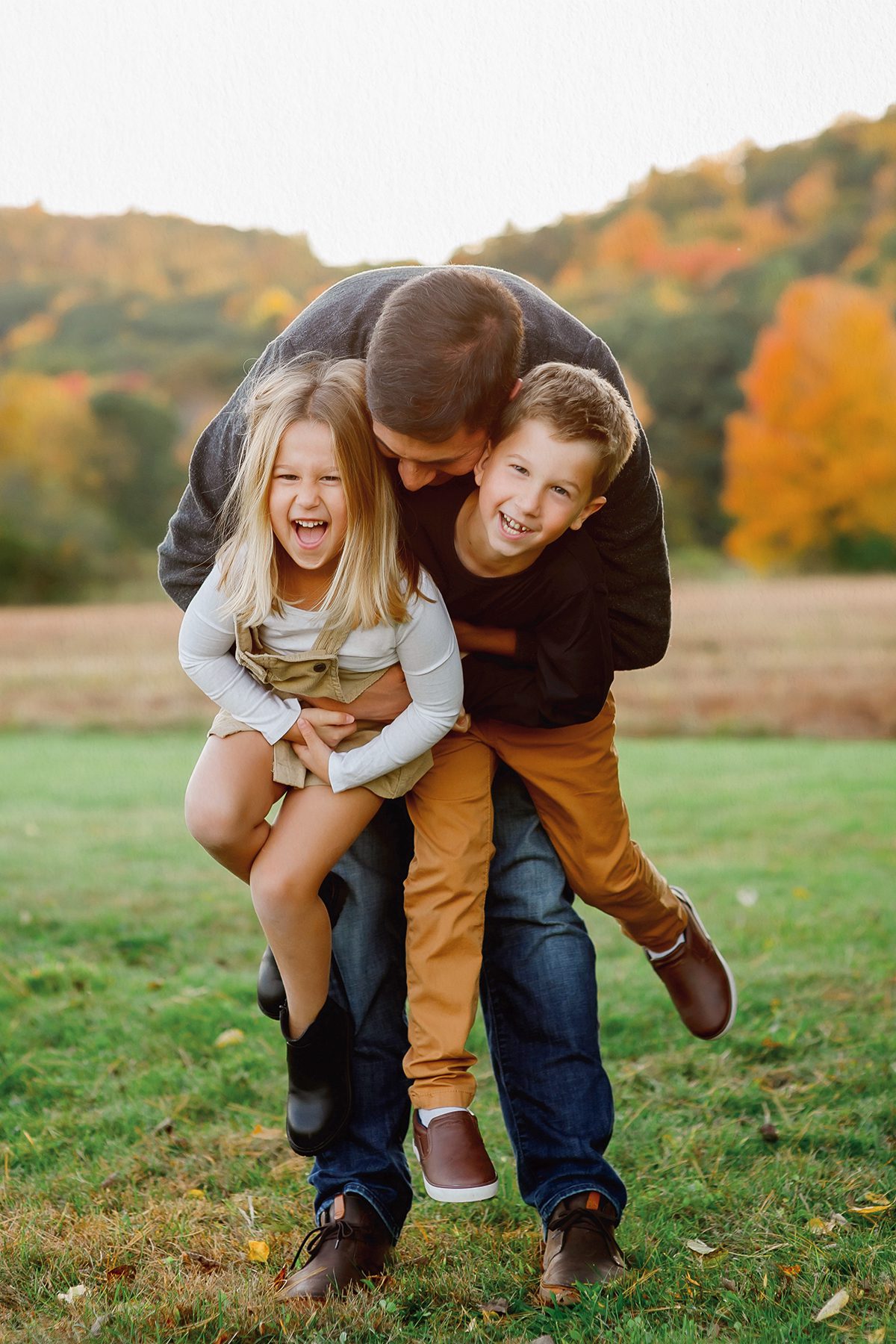 A family photoshoot featuring a father playing with his two children in an open field in the fall.
