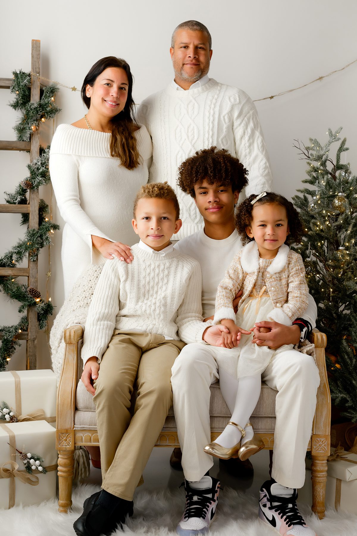 Family of five smiling together in front of a neutral holiday backdrop with twinkling lights and greenery.