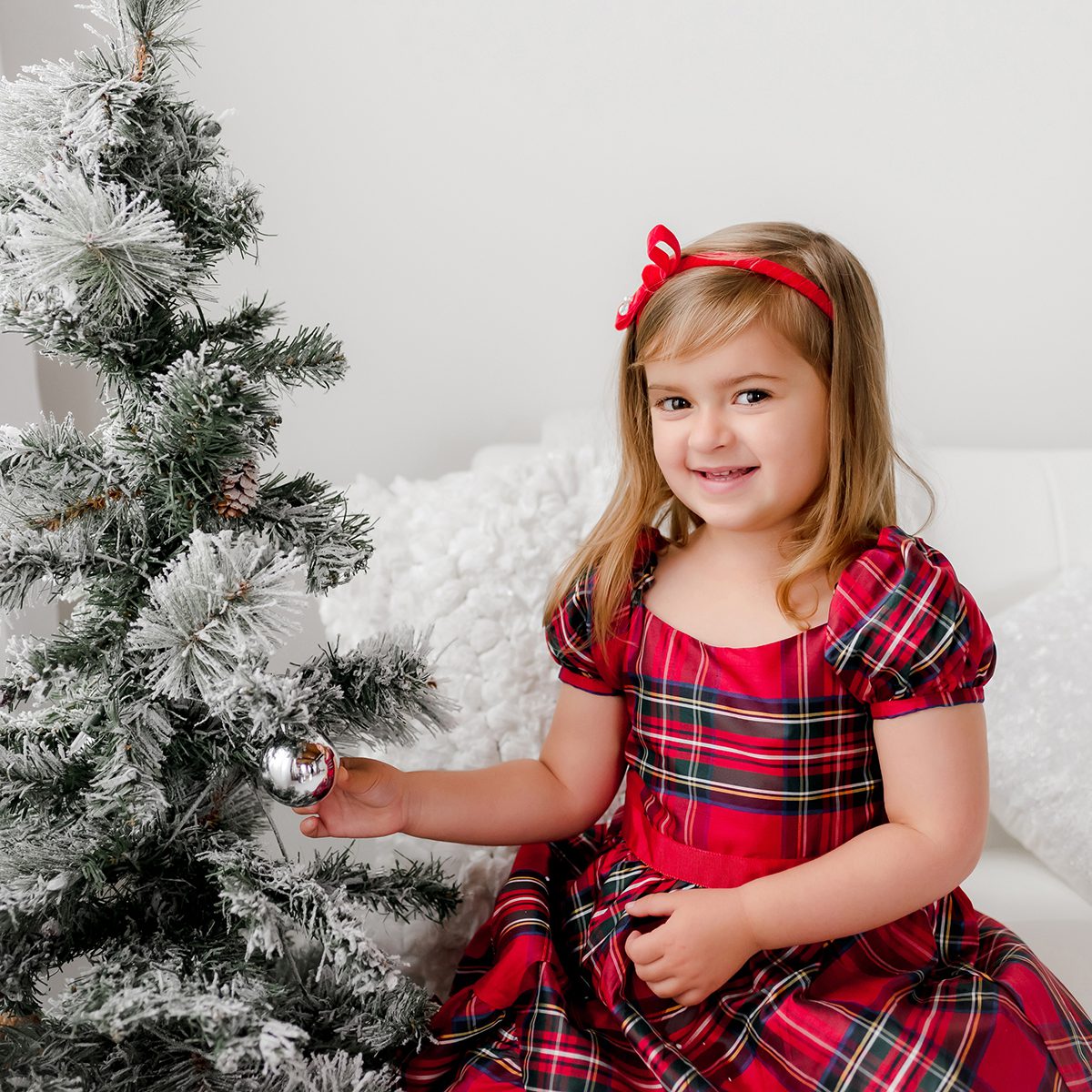 12_SANTA_CHRISTMAS_PHOTOS Little girl sitting next to a Christmas tree for a traditional holiday portrait. She is wearing a red plaid dress. Taken at Kelli Dease Photography during a Christmas mini session in Farmington, Connecticut.