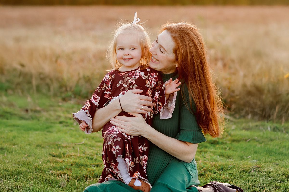 A mom and daughter in an open field in fall, surrounded by autumn colors and soft golden hour light.