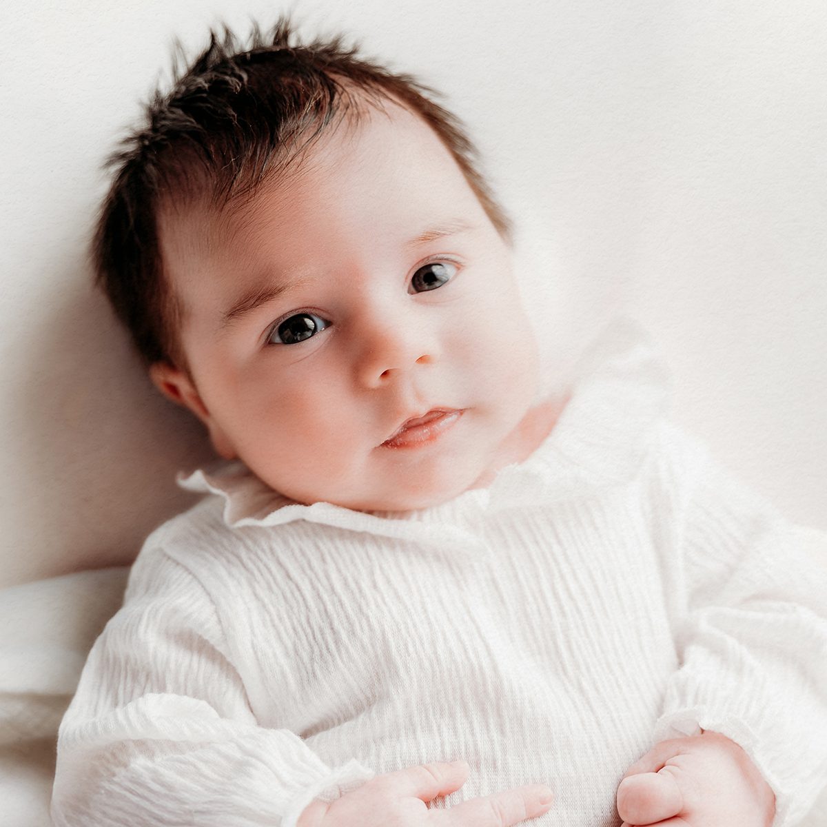 Studio baby portrait of an alert infant with dark hair and bright eyes, wearing a soft white outfit and lying against a light neutral background during a minimalist baby photography session.