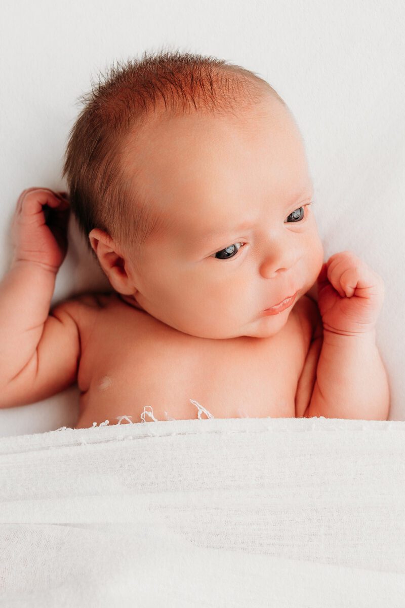 Studio newborn portrait of an alert baby lying on their back with bright eyes and hands raised, photographed against a clean white background during a newborn session.