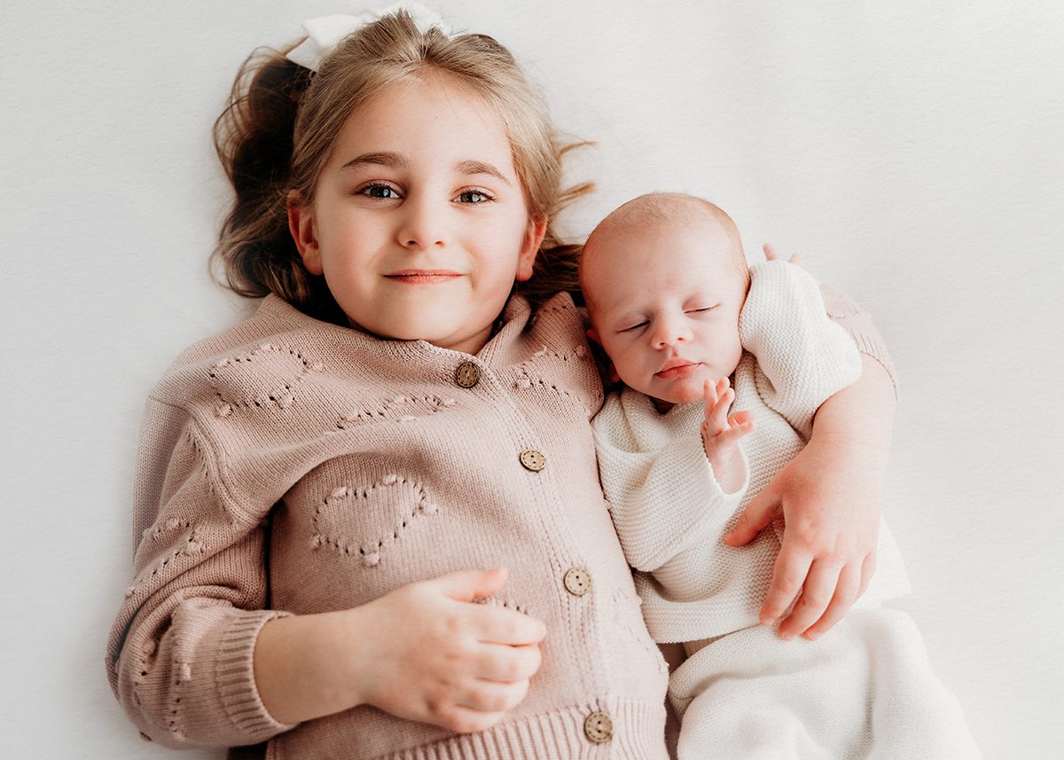 Studio sibling portrait of an older sister lying beside her newborn sibling wrapped in white, photographed against a soft neutral background