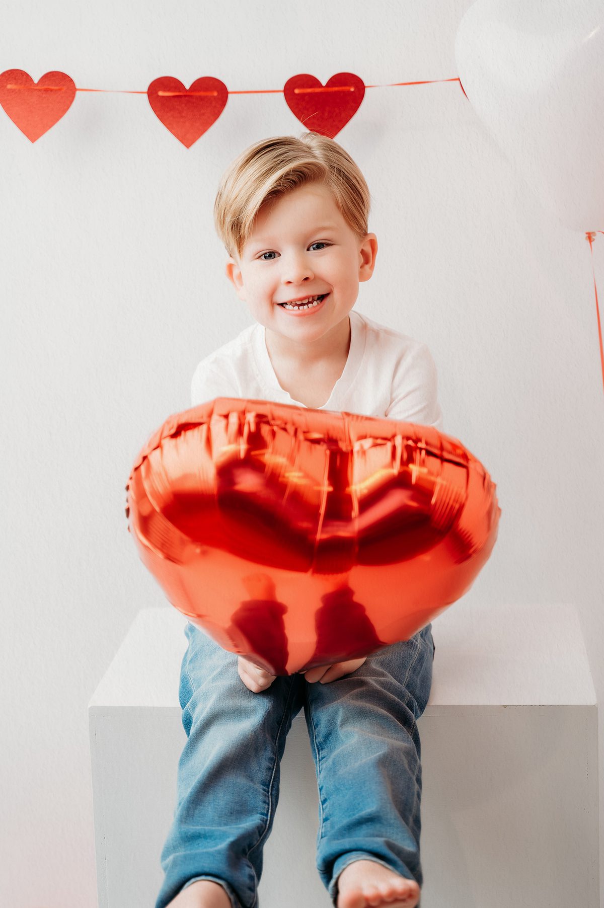 Valentine-themed studio photo of a 4 year old boy surrounded by red heart balloons and natural light