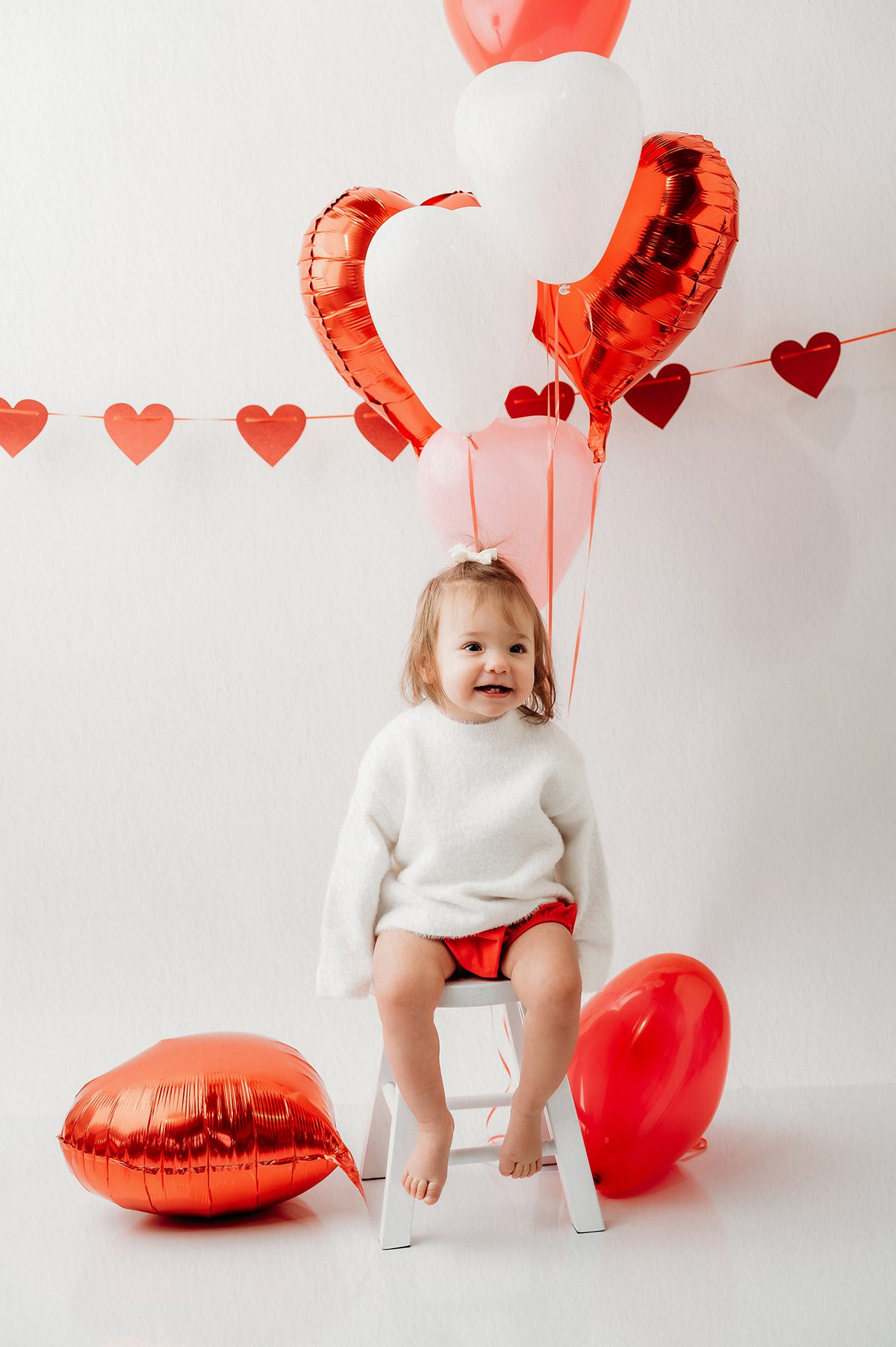 Valentine mini session portrait of a young child surrounded by red heart balloons in a bright, clean studio setting in Connecticut.