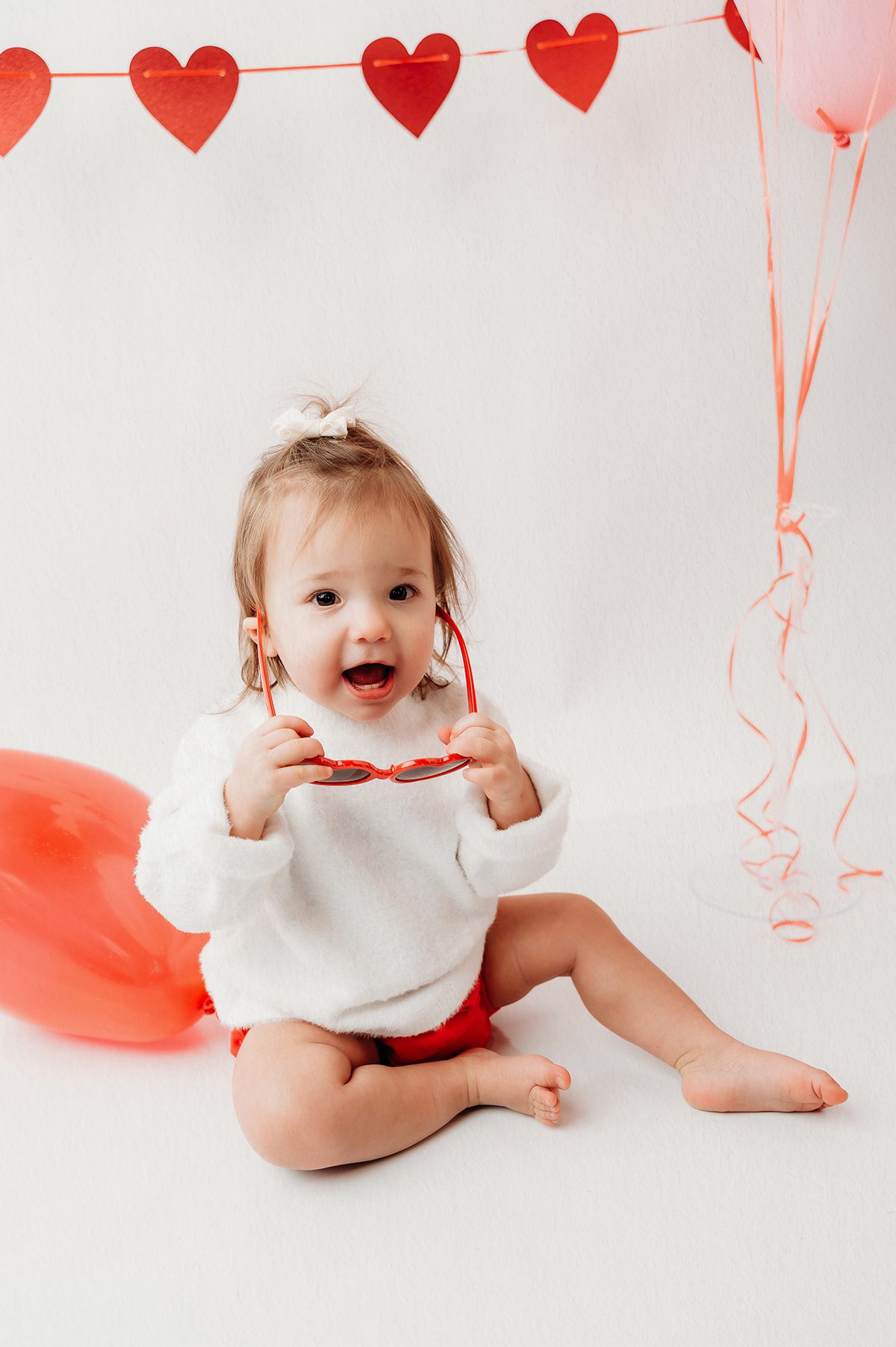 Valentine mini session photo of a baby wearing soft neutral clothing with red heart balloons in a Connecticut photography studio.
