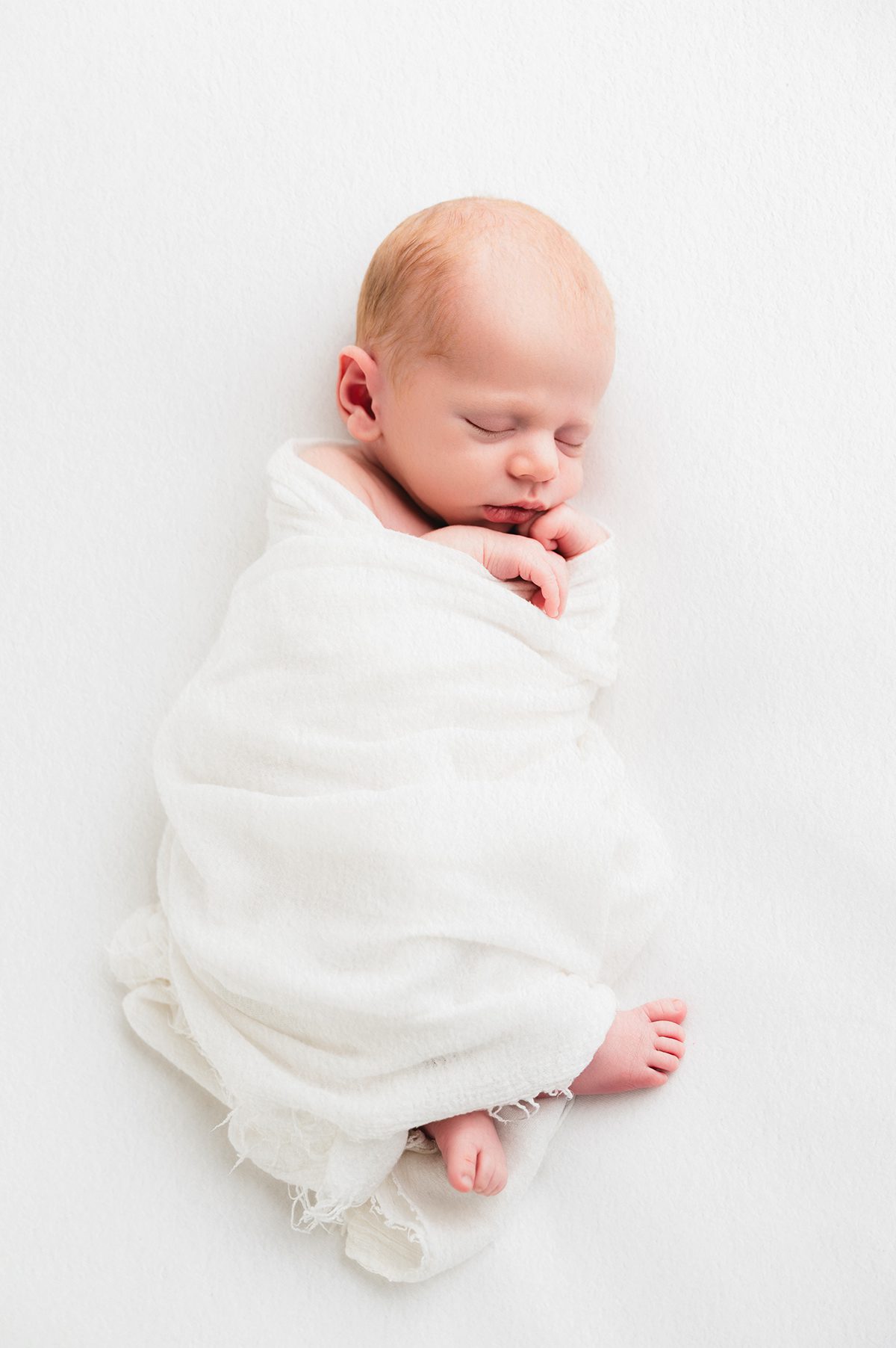 Sleeping newborn baby swaddled in soft white fabric, curled on a clean white backdrop during a minimalist Connecticut studio newborn photography session.