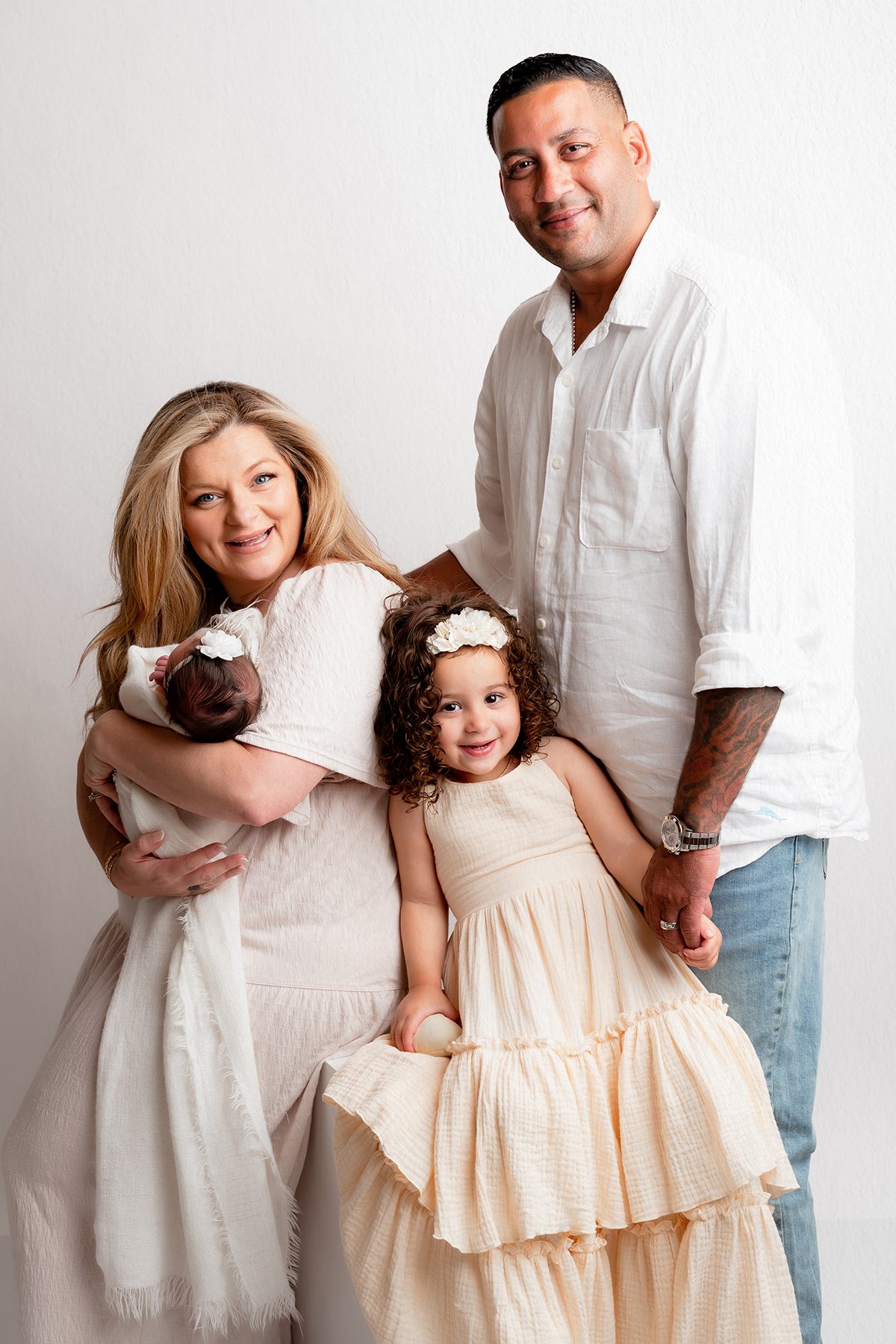 A family of four dressed in neutral tones poses together during a studio newborn photoshoot, showing coordinated outfits for parents, a toddler, and a newborn.