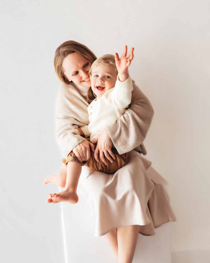 Mom in beige sweater sitting and hugging laughing toddler boy during a Connecticut studio motherhood mini session