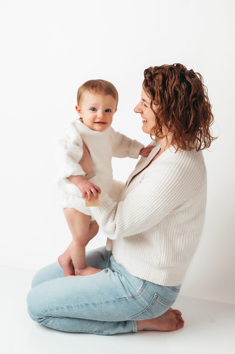 Mom sitting cross-legged on studio floor with baby standing up and smiling during a Connecticut motherhood mini session