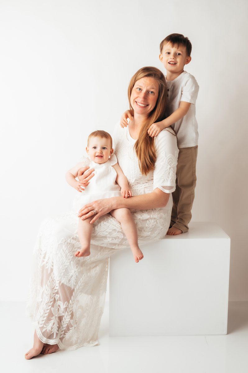 Mom in white lace dress seated on cube with baby in her lap and young son standing behind her during a Connecticut motherhood mini session