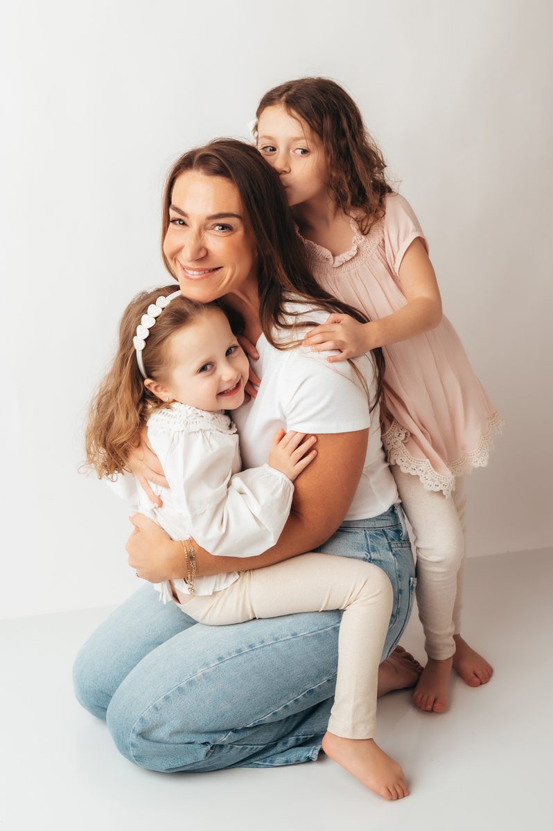 Mom sitting on studio floor with two daughters hugging her from both sides during a Connecticut motherhood mini session