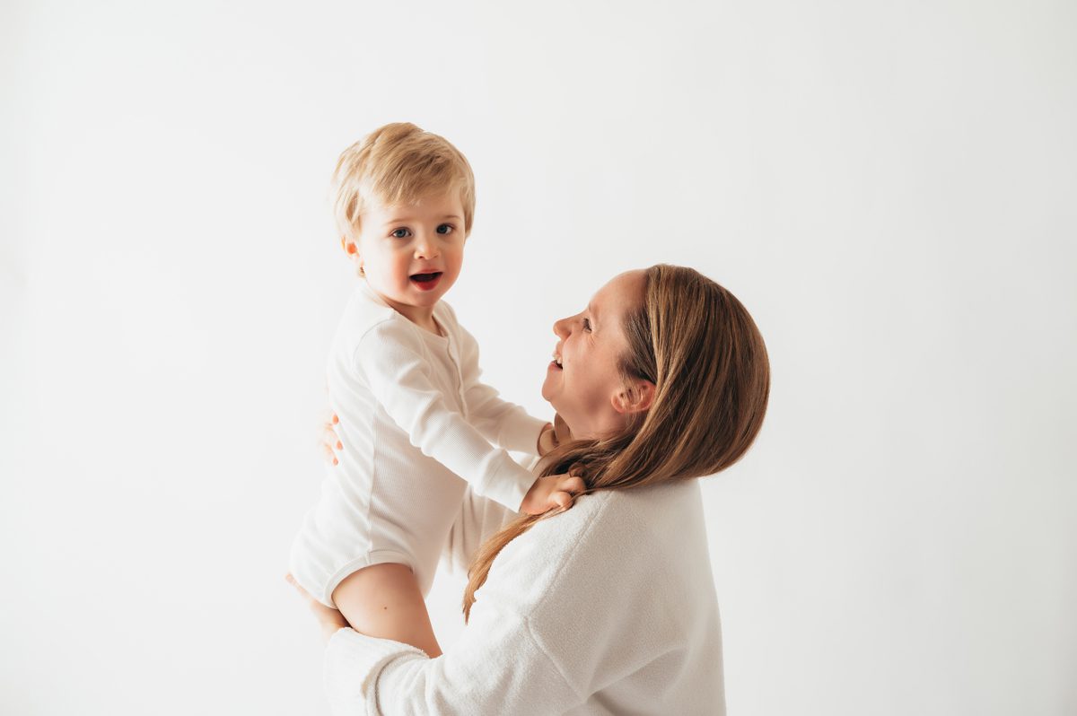 Mother lifting laughing blonde toddler boy overhead while both smile at each other during a Connecticut motherhood mini session