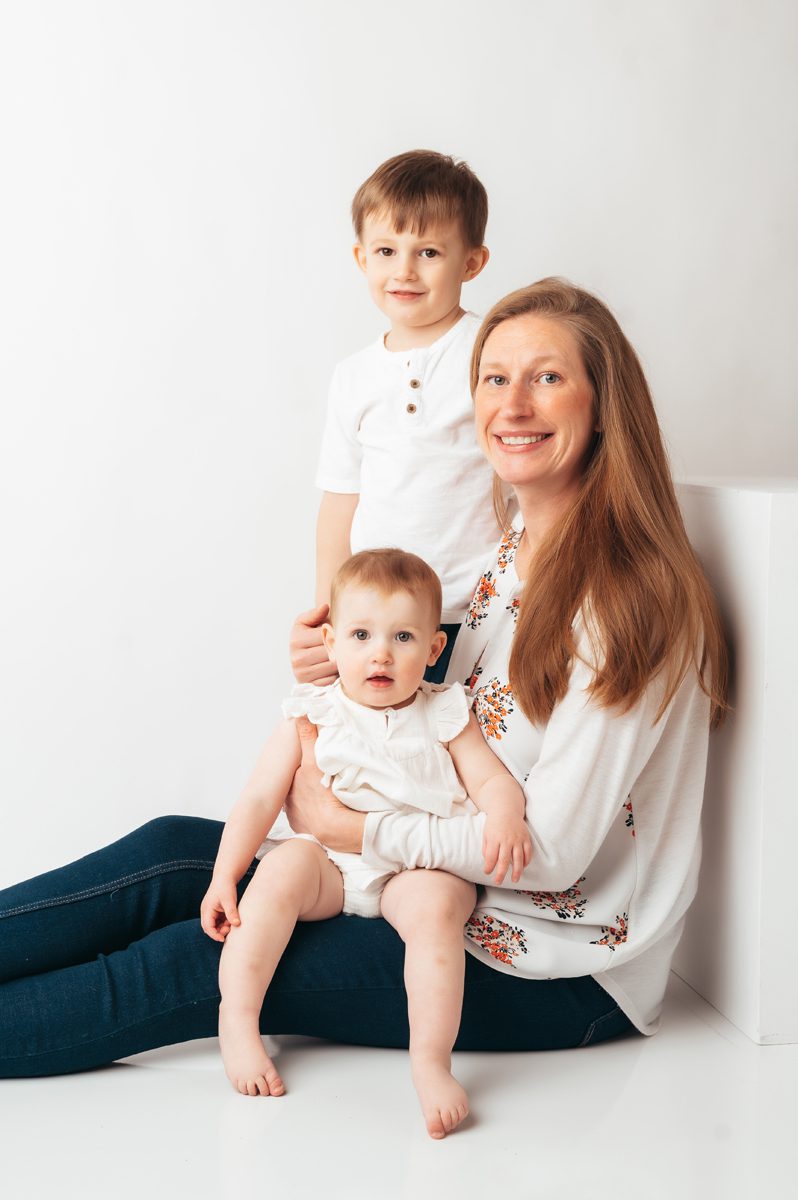 Mom seated on studio floor smiling with baby on her lap and young son standing beside her during a Connecticut motherhood mini session