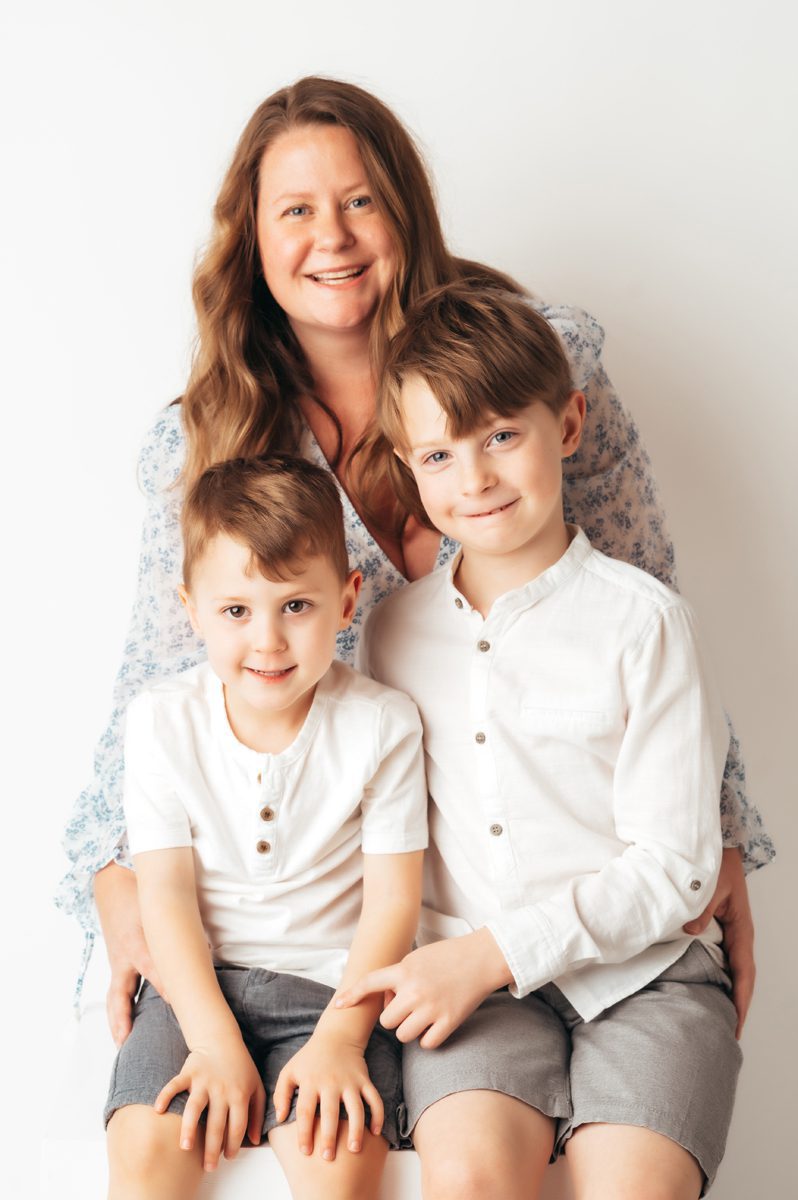 Mom in blue floral dress seated behind two young sons smiling at camera during a Connecticut motherhood mini session