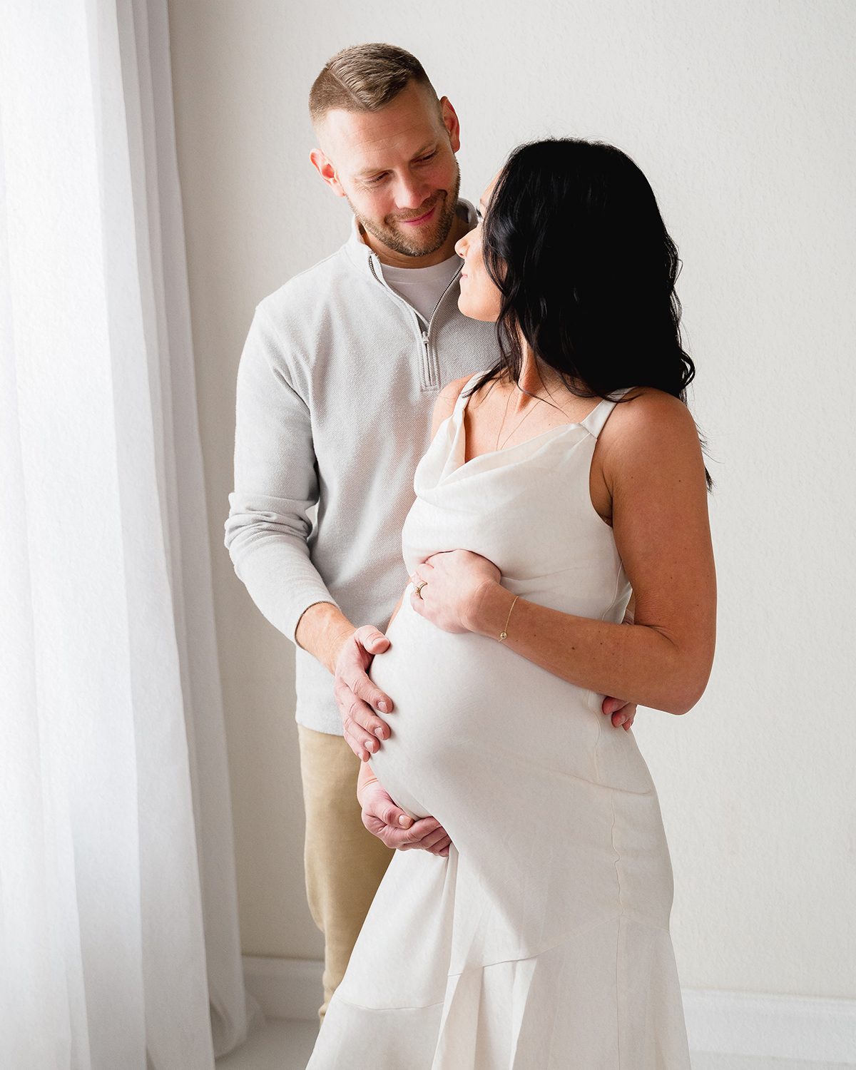 Expecting couple wearing neutral outfits, photographed against a soft studio backdrop during a maternity session at Kelli Dease Photography