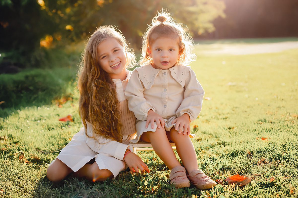 Two young sisters in cream and white outfits standing in a sunlit field during a family photo session, photographed by Kelli Dease Photography in Farmington, CT.