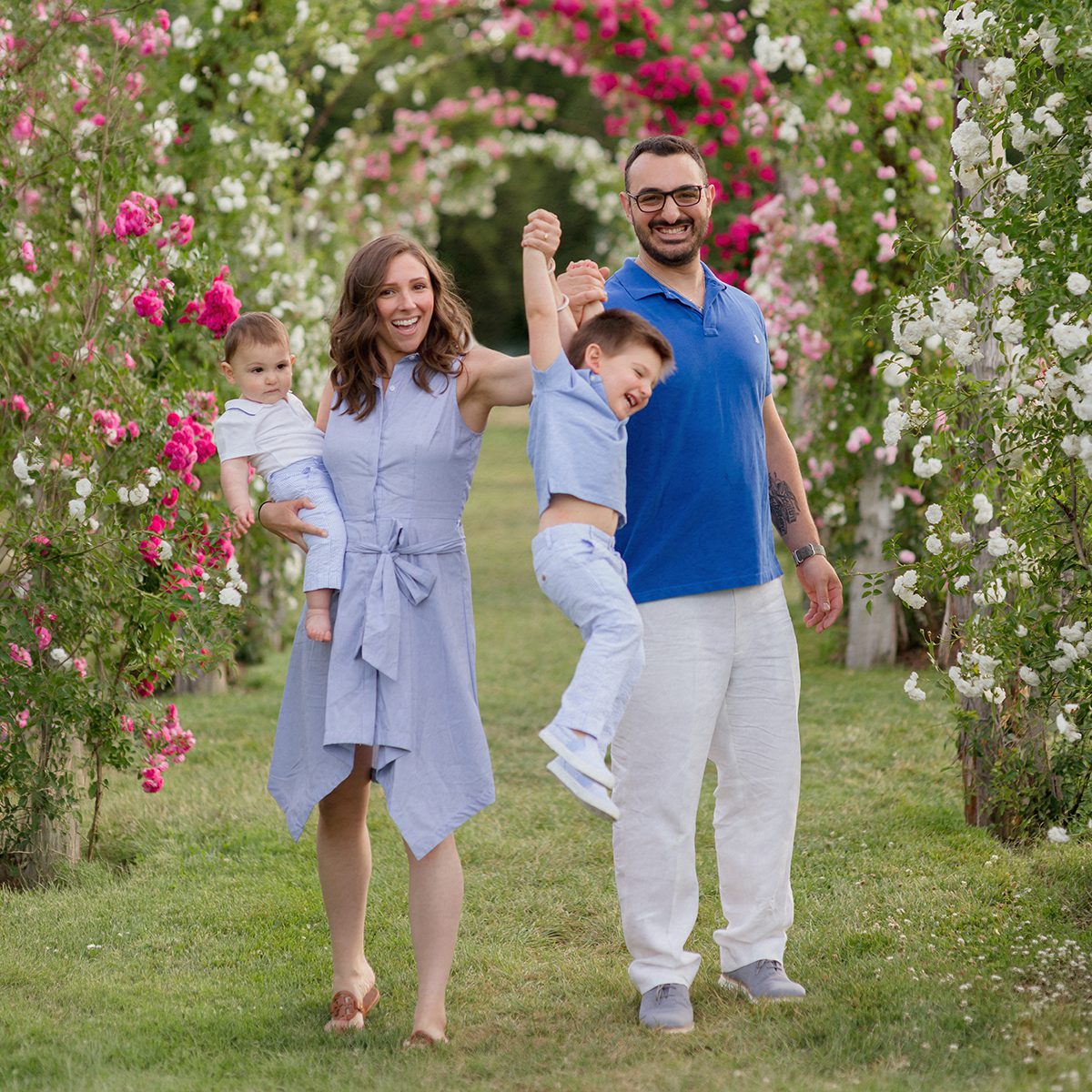 A family of four walks through the rose garden at Elizabeth Park in Hartford, CT. The mom holds a baby on her hip while the dad swings their laughing toddler by one arm. Both parents are smiling. They're dressed in coordinating shades of blue and white, surrounded by arched trellises covered in pink and white blooming roses.