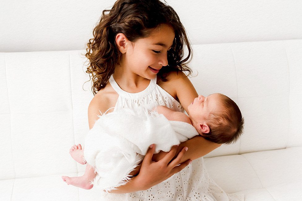 Young girl in a white eyelet dress holding her newborn sibling on a white couch during a sibling newborn session at Kelli Dease Photography in Farmington, CT