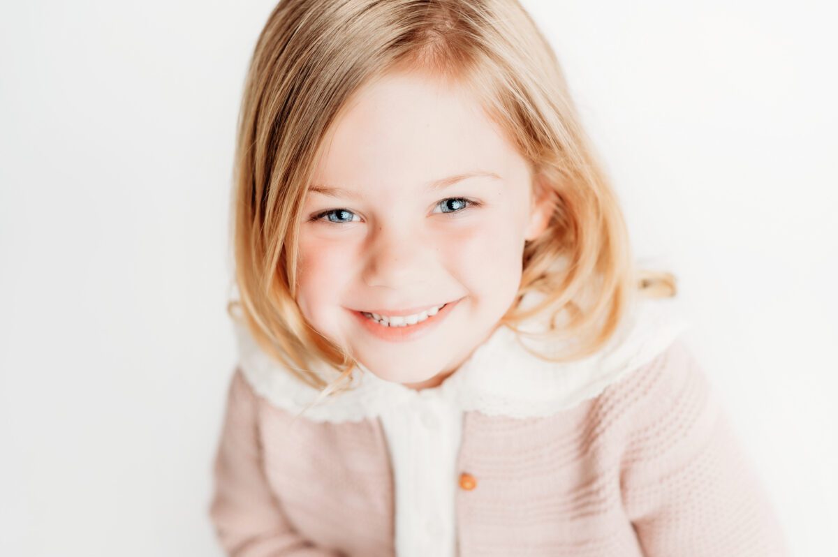 a head and shoulders image of a five year-old girl in a pink sweater, smiling during a personality portrait photoshoot in farmington, ct.