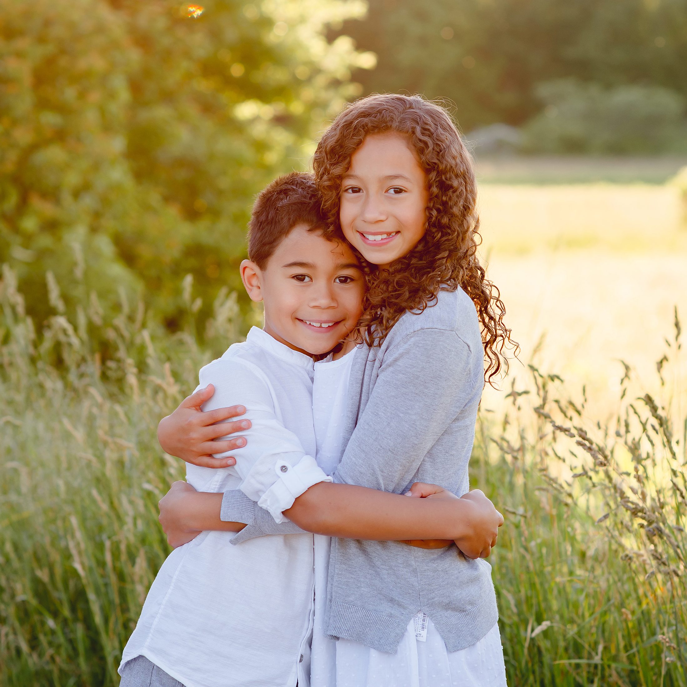 A girl with long curly hair hugs her younger brother from behind in a sun-drenched meadow in Simsbury, CT. Both children are smiling at the camera. She's wearing a gray cardigan and he's in a white button-down shirt.