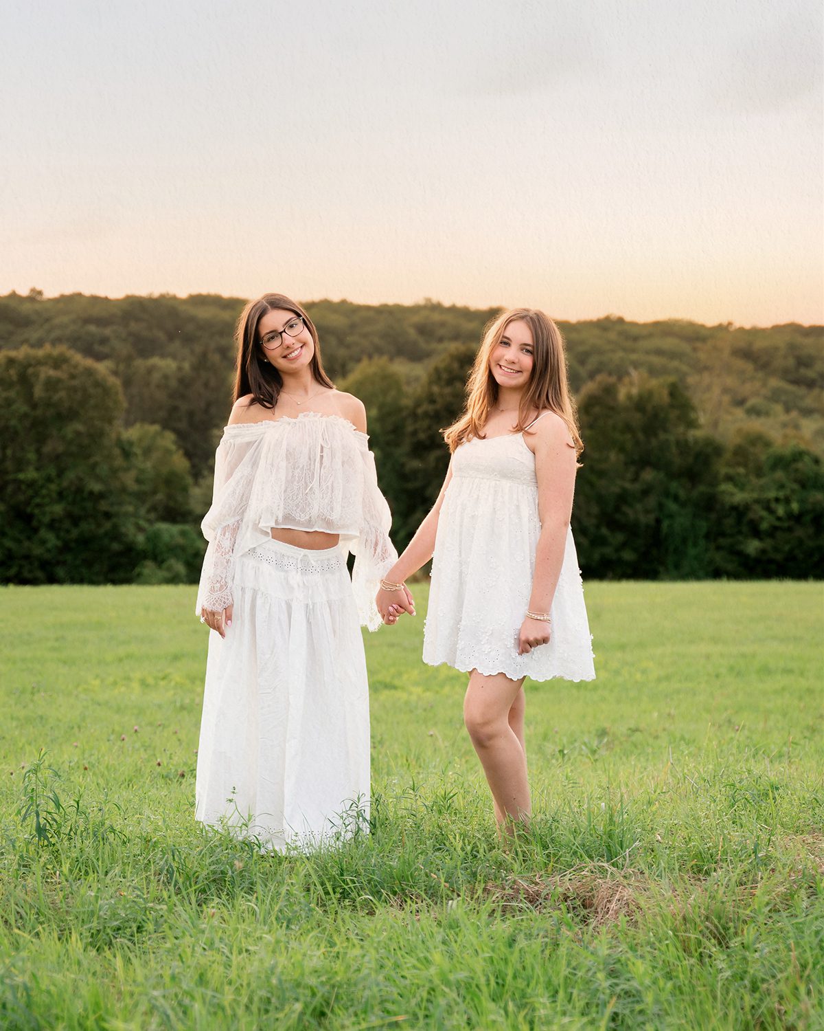 Two teenage sisters standing in a field during sunset, holding hands and wearing bohemian white dresses. Photographed by Kelli Dease in Litchfield, Connecticut.