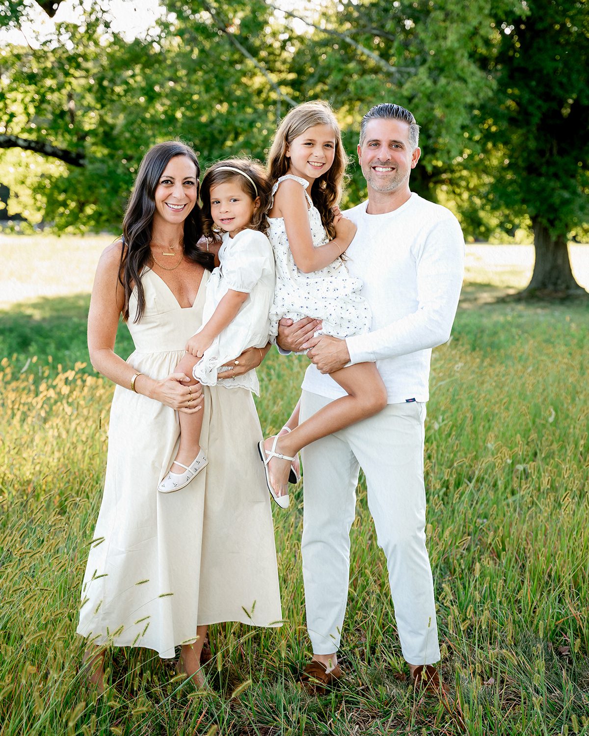 A family of four with two young girls standing in a field at Waveney Park in New Canaan, Connecticut. The family is wearing neutrally colored clothing and smiling at the photographer, Kelli Dease.