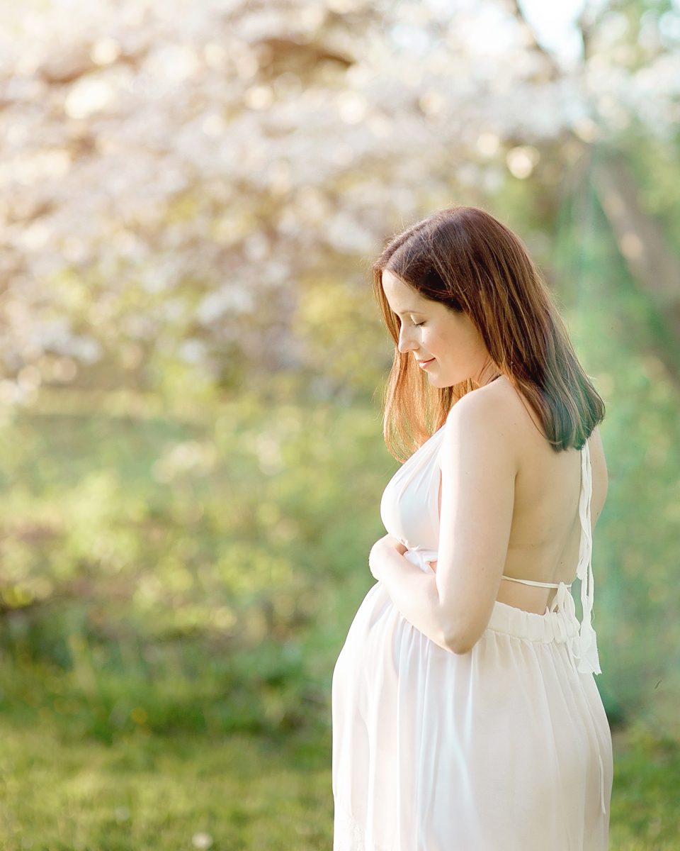 Pregnant woman in a white dress cradling her bump among flowering trees during a spring maternity session at Elizabeth Park in West Hartford