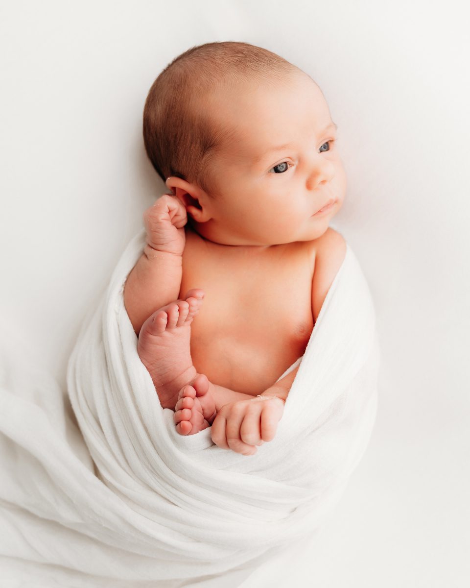 Wide-awake newborn wrapped in white with one foot peeking out, looking toward the camera during a studio newborn session