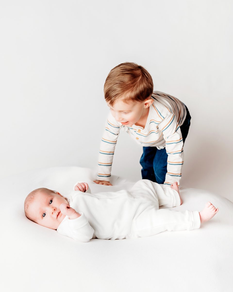 Older brother leaning over his newborn sibling on a white backdrop during a studio newborn session