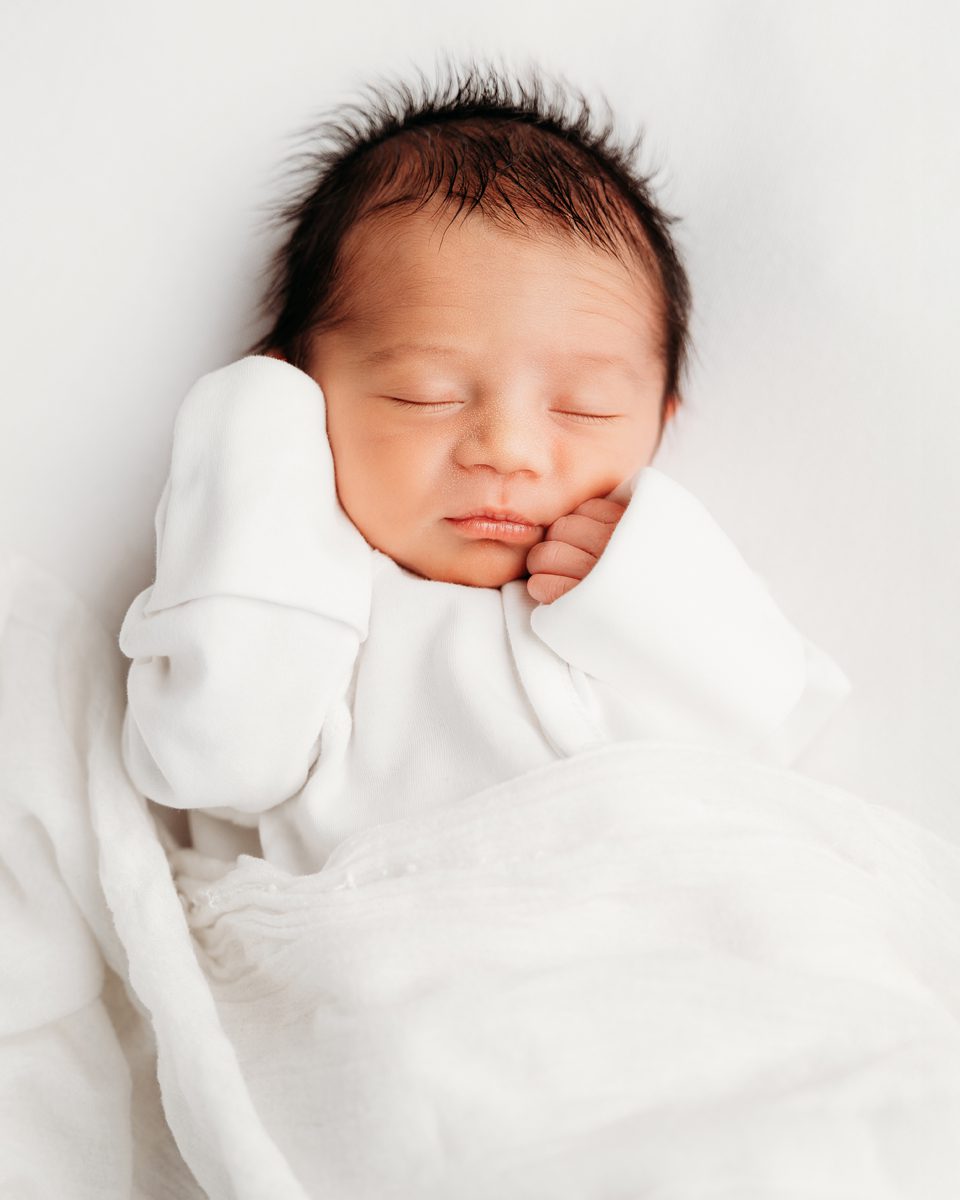 Dark-haired newborn sleeping with hands tucked under the chin, swaddled in white, during a West Hartford area newborn session