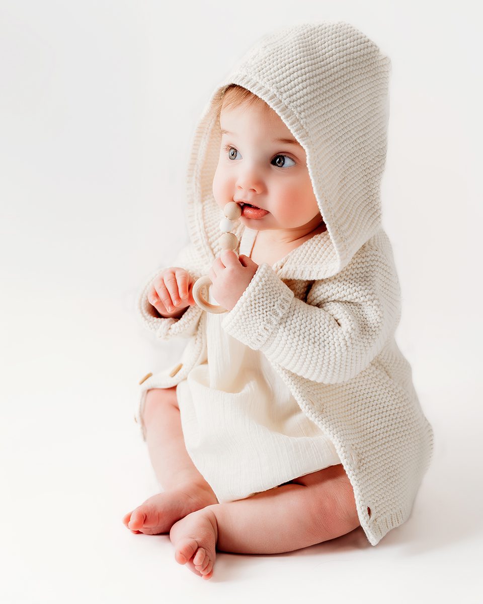 a baby girl dressed in a hooded cream sweater, biting on a toy, during a milestone photo session with Connecticut photographer Kelli Dease