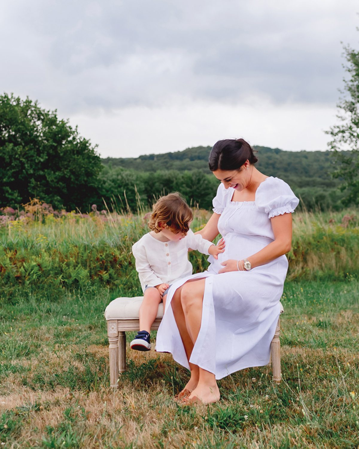 farmington ct outdoor maternity photography_02 SQ Expectant mother in a white dress seated on a vintage bench in a grassy field in Simsbury, CT, her toddler son leaning in to touch her belly, outdoor maternity session at Kelli Dease Photography