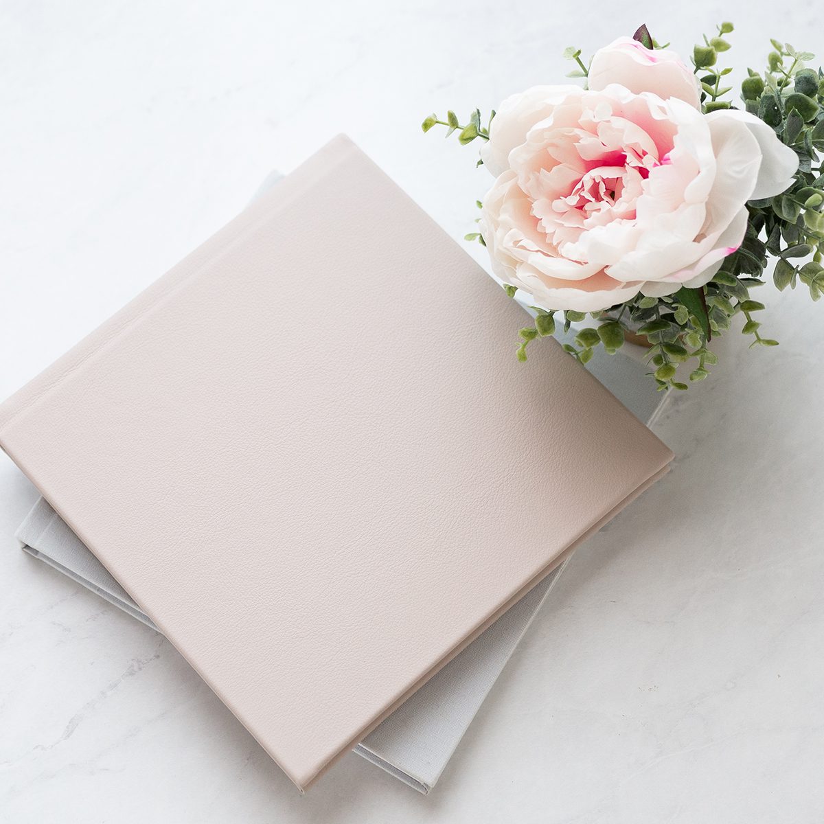 a photo album with a pink blush leather cover on a marble table with a pink peony in the background